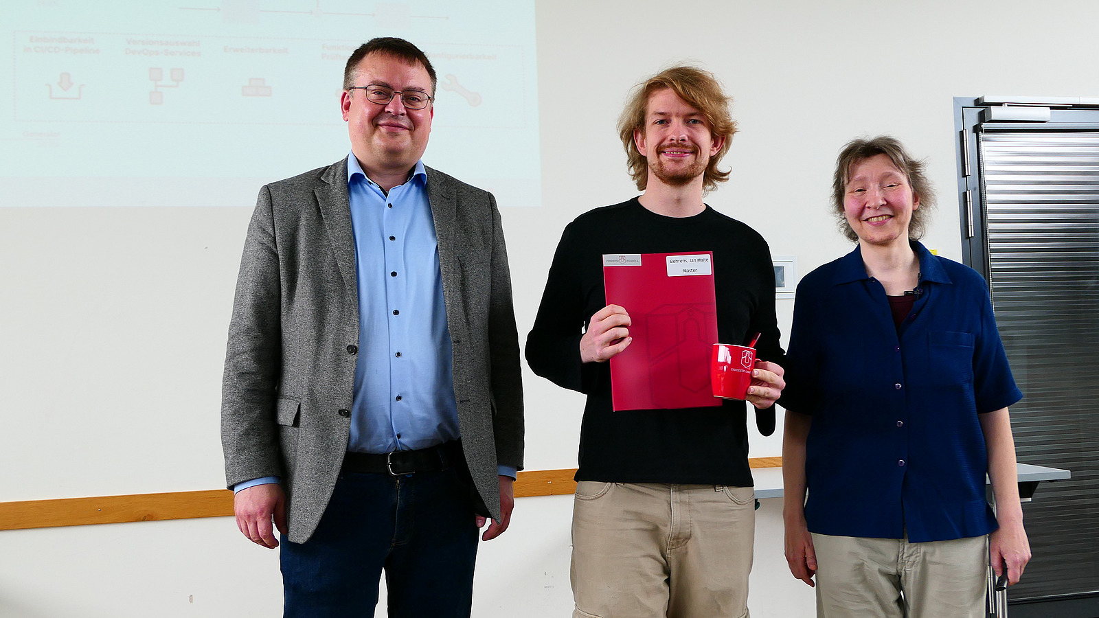 Ein Student mit Studiendekanin und Dekan stehen nebeneinander. Der Student hat eine Mappe in der Hand