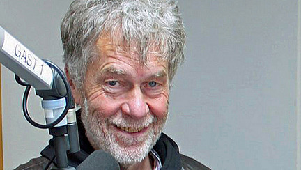 Ein älterer Mann mit grauem, zerzaustem Haar und einem freundlichen Lächeln steht vor einem Mikrofon. Er trägt eine dunkle Jacke. Der Hintergrund ist neutral und weist auf eine Studioatmosphäre hin.
