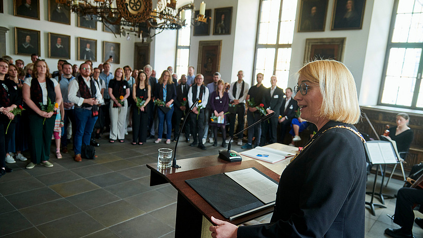 In a historical-looking room, a person stands at a lectern in the foreground. Numerous people are standing in front of her and looking at her.