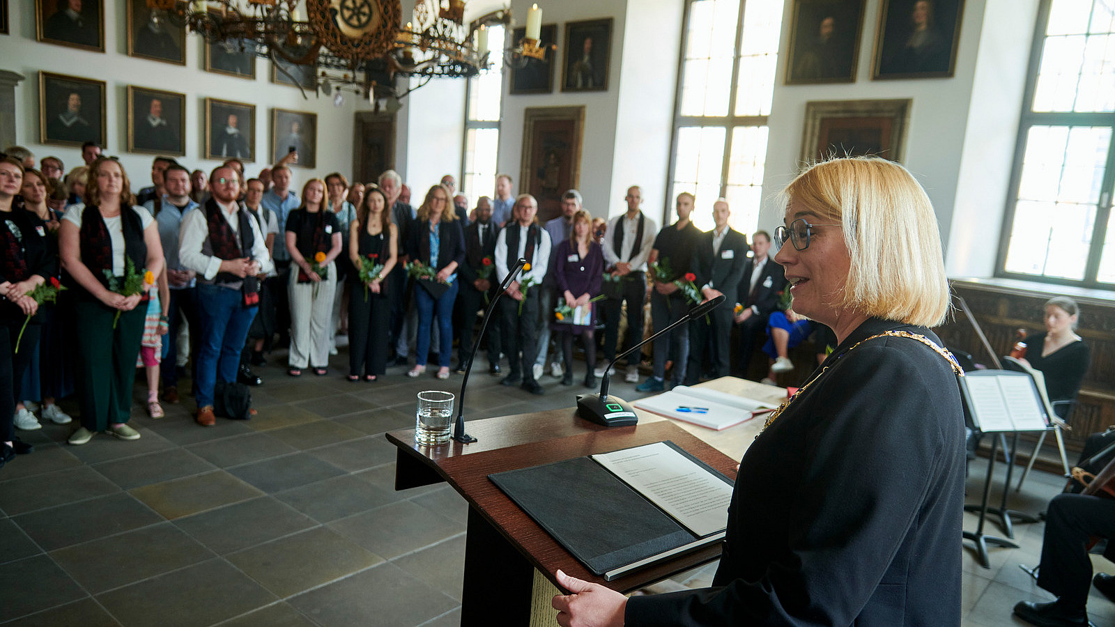 In a historical-looking room, a person stands at a lectern in the foreground. Numerous people are standing in front of her and looking at her.