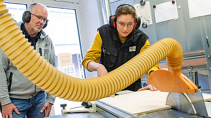 Eine weibliche Person schiebt mit konzentriertem Blick ein Holzbrett durch eine Kreiss&auml;ge. Neben ihr steht der Ausbilder und schaut zu.