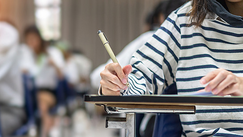 In a classroom, several children sit at their desks, hands raised to sign in. A boy in a checked shirt and a girl in a pink T-shirt are clearly visible. In the foreground, the teacher, who is standing at a blackboard and addressing the pupils, looks in their direction.