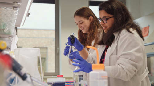 Two women wearing lab coats and blue gloves in a laboratory, the one in front holding a pipette. 