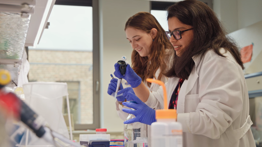 Zwei Frauen mit Kittel und blauen Handschuhen im Labor, die vordere h&auml;lt eine Pipette. 
