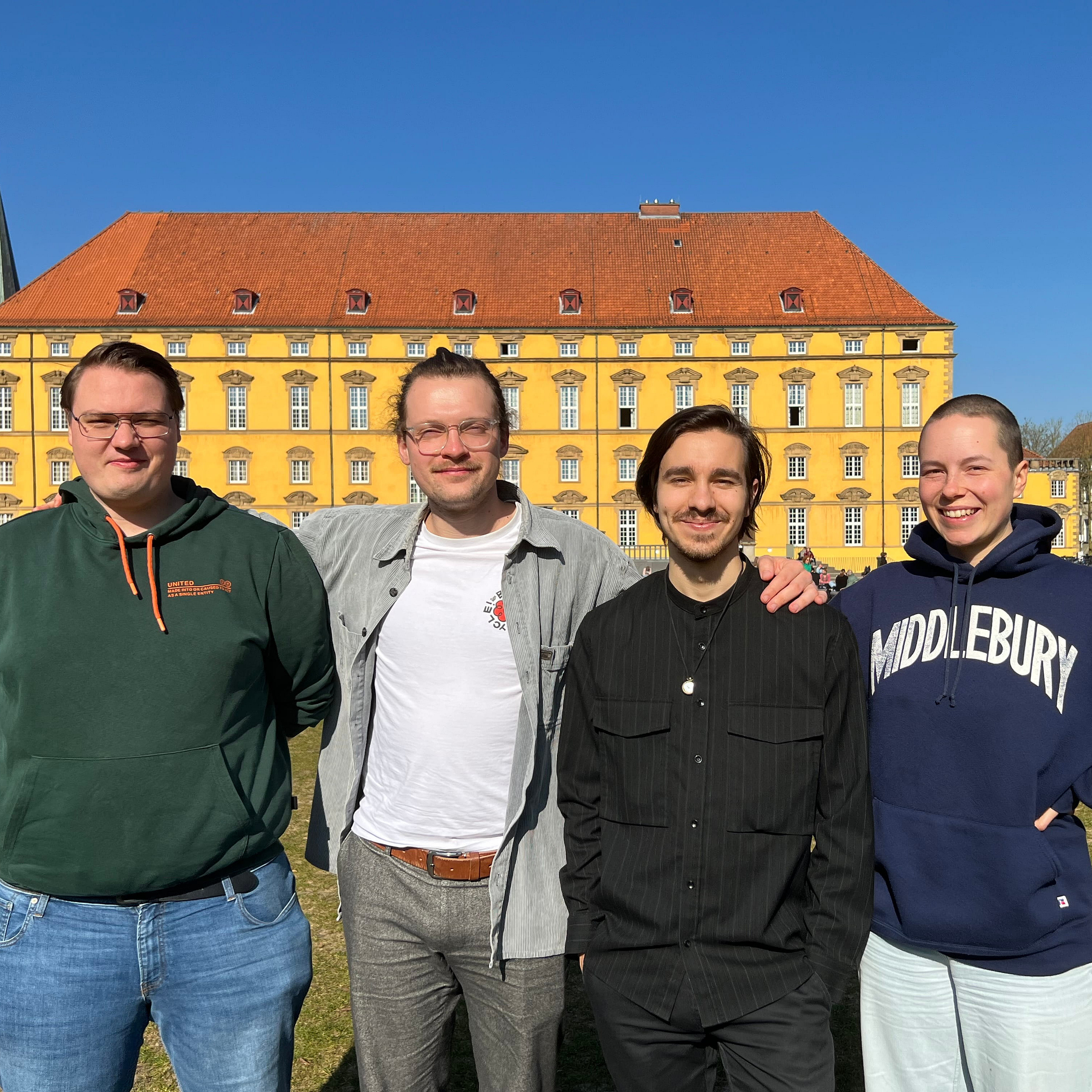 Four students from the Student Board are standing side by side in the sun, looking ahead. In the background, you can see the yellow university castle and the green castle gardens.