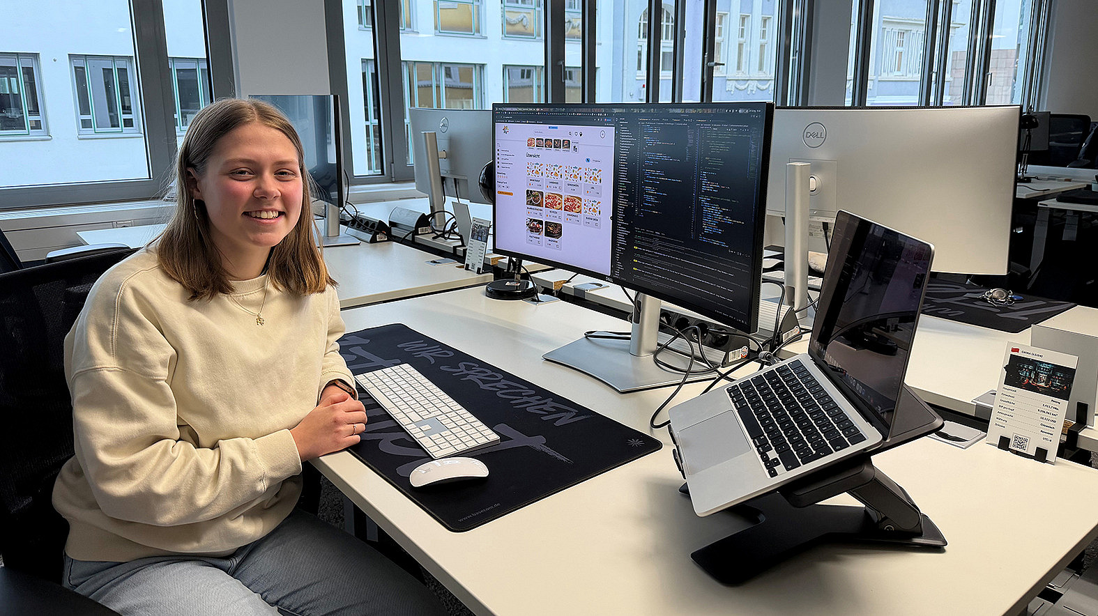Eine Frau sitzt an einem Laptop in einem hellen Raum und arbeitet. Im Hintergrund sind Fenster zu erkennen.
