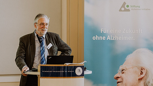 Roland Brandt stands at a lectern, next to him is a poster with the words ‘Für eine Zukunft ohne Alzheimer’.