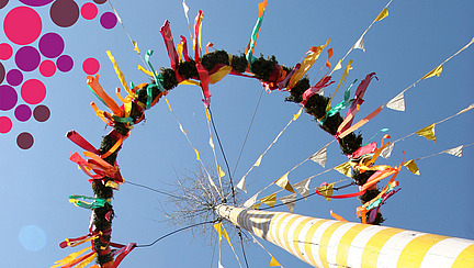 View from below of the top of a maypole with colourful fluttering ribbons on a green wreath. In the background blue sky, in two corners of the picture there are colourful confetti circles as a frame.