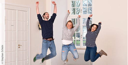 Ein junger Mann und zwei junge Frauen springen erfreut in die Luft und reißen die Arme nach oben