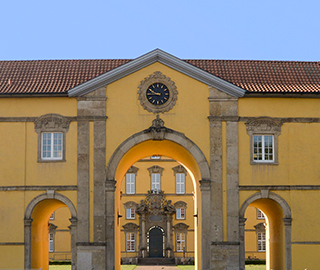 Das Haupttor des Schlosses über eine Straße fotografiert. Über dem Tor ist eine Uhr in die Mauer eingelassen.