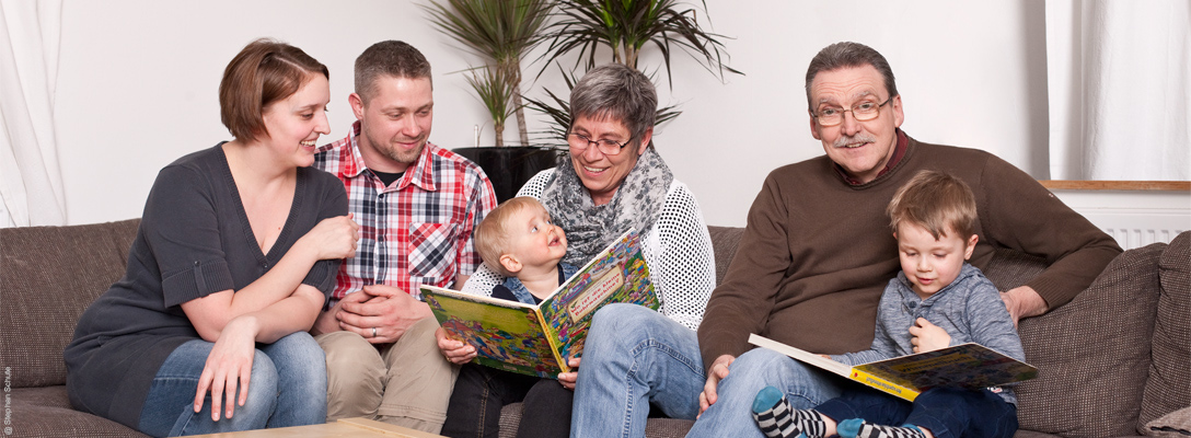 Eine Familie mit mehreren Generationen sitzt gemeinsam auf einem Sofa. Die beiden Kinder schauen auf dem Schoß ihrer Großeltern Bücher mit ihnen an. Die Eltern sitzen daneben und blicken auf das Geschehen. Foto: Stephan Schute