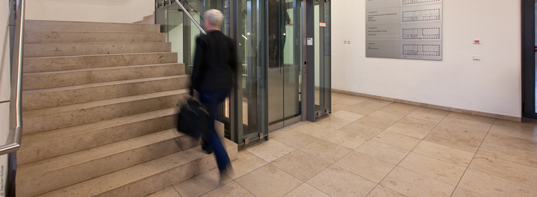 Ein Mann mit einer Aktentasche geht in einem Universitätsgebäude eine Treppe hinauf. Er ist dabei nur verschwommen zu sehen und befindet sich demnach in Bewegung. Foto: Stephan Schute