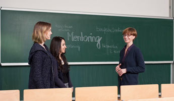 Das Bild zeigt zwei junge Frauen mit einer Mentorin. Sie stehen vor einer Tafel der Universität. Auf der Tafel ist groß "Mentoring" zu lesen. Um den Begriff herum steht: "Kompetenzen erweitern", "berufliche Orientierung", "Netzwerken und Kontakte knüpfen" und  "Karriereplanung". Foto: Stephan Schute