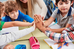 Kinder und Lehrer sitzen an einem Tisch und führen die Hände zusammen.