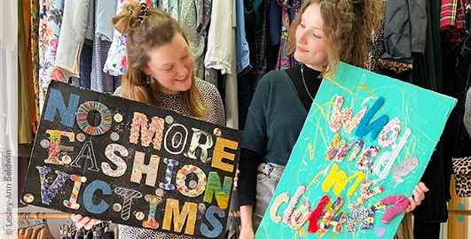 Zwei junge Frauen vor Regalen mit Kleidung, sie halten Plakate in den Händen mit Aufschrift "No more fashion victims" und "Sho made my clothes?"