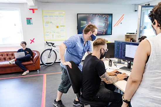 StartUp-Athmosphäre, in einem großen Arbeitsraum mit Personen am Rechner, einer Person auf dem Sofa mit Laptop, ein Rennrad steht an der Wand