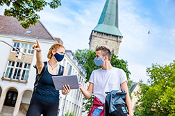 Zwei Studierende mit Alltagsmasken stehen vor einem Uni-Gebäude. Die Frau zeigt dem Mann etwas auf ihrem Laptop