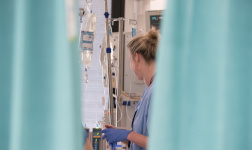 Eine weibliche Pflegekraft arbeitet mit Hanschuhen an einem  Krankenhausbett, im Vordergrund ist ein Vorhang zu sehen