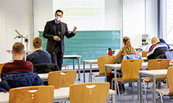 Studierende in Seminar, Lehrender mit Schutzmaske steht vorne und redet
