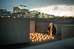 Leuchtende Kerzen zwischen den Denkmal-Stelen, eine Frau kniet davor und zündet ein weiteres Licht an