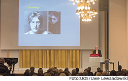 [Translate to English:] Redner Heinrich Detering auf der Bühne, im Hintergrund Fotos von John Lennon