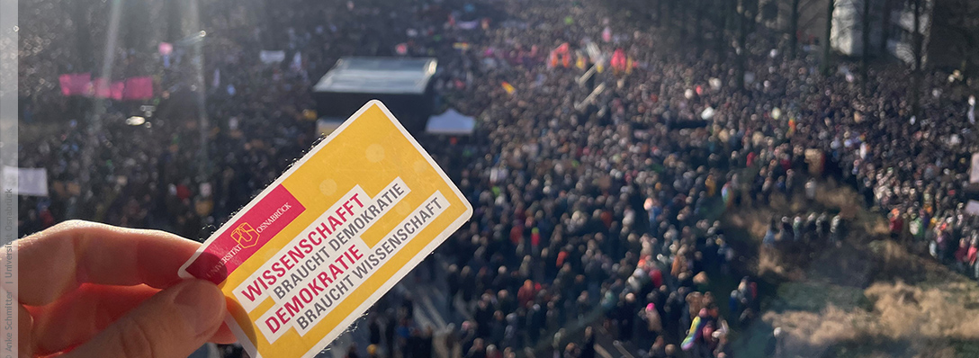 Im Hintergrund demonstrierende Menschen, vorne Aufnäher mit Aufschrift "Wissenschaft braucht Demokratie, Demokratie braucht Wissenschaft"