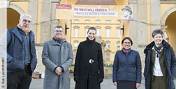 Referent*innen stehen vor einem Remarque-Zitat-Banner ("Die Welt will Frieden trotz gewisser Politiker") am Torbogen des Osnabrücker Schlosses