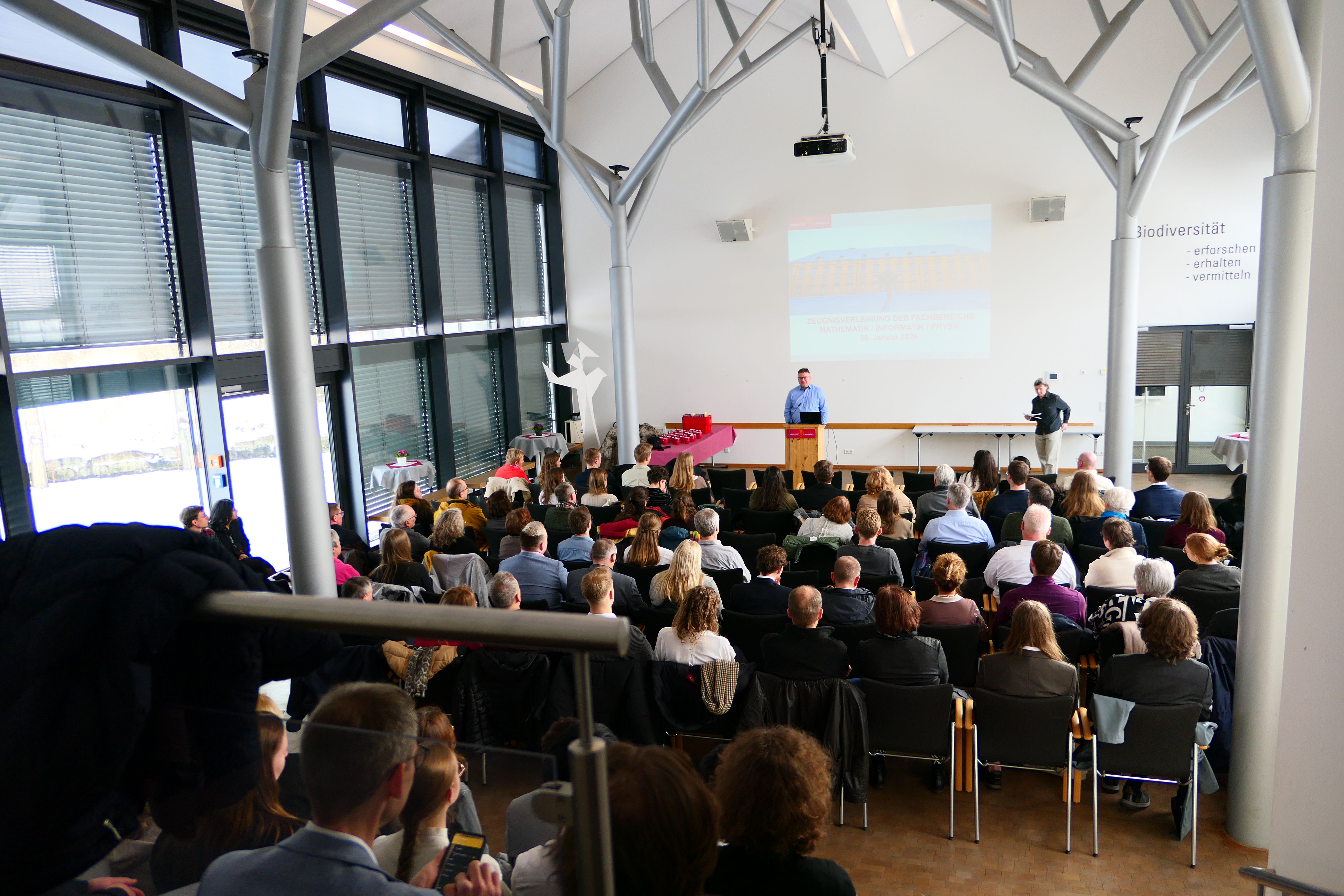 2 Personen stehen vor etwa 100 sitzenden Personen.