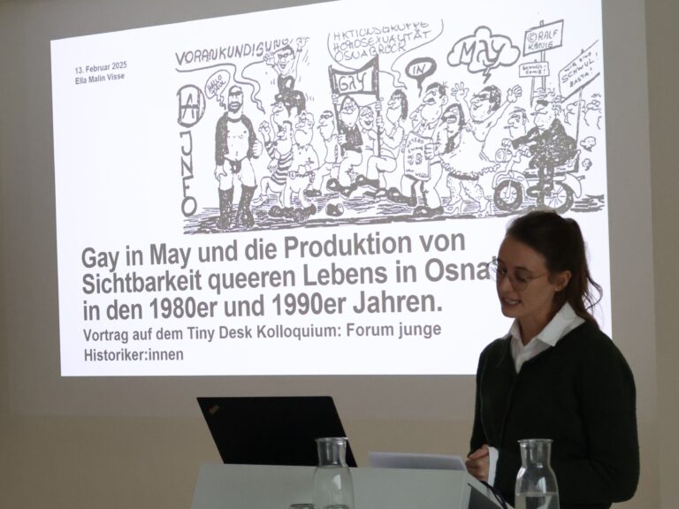 Eine Frau mit dunkelblonden Haaren und Brile in einem dunklen Pulloversteht liest von ihrem Skript ab und hält einen Vortrag, zu dem eine Präsentation mit einem schwarz-weißen Bild an die Wand hinter ihr projiziert wird.