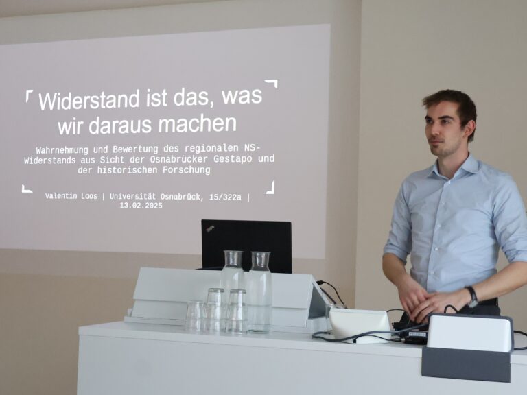 Ein Mann mit dunklen Haaren und hellem Hemd steht hinter einem Tisch und hält einen Vortrag, zu dem eine Präsentation an die Wand hinter ihm projiziert wird.