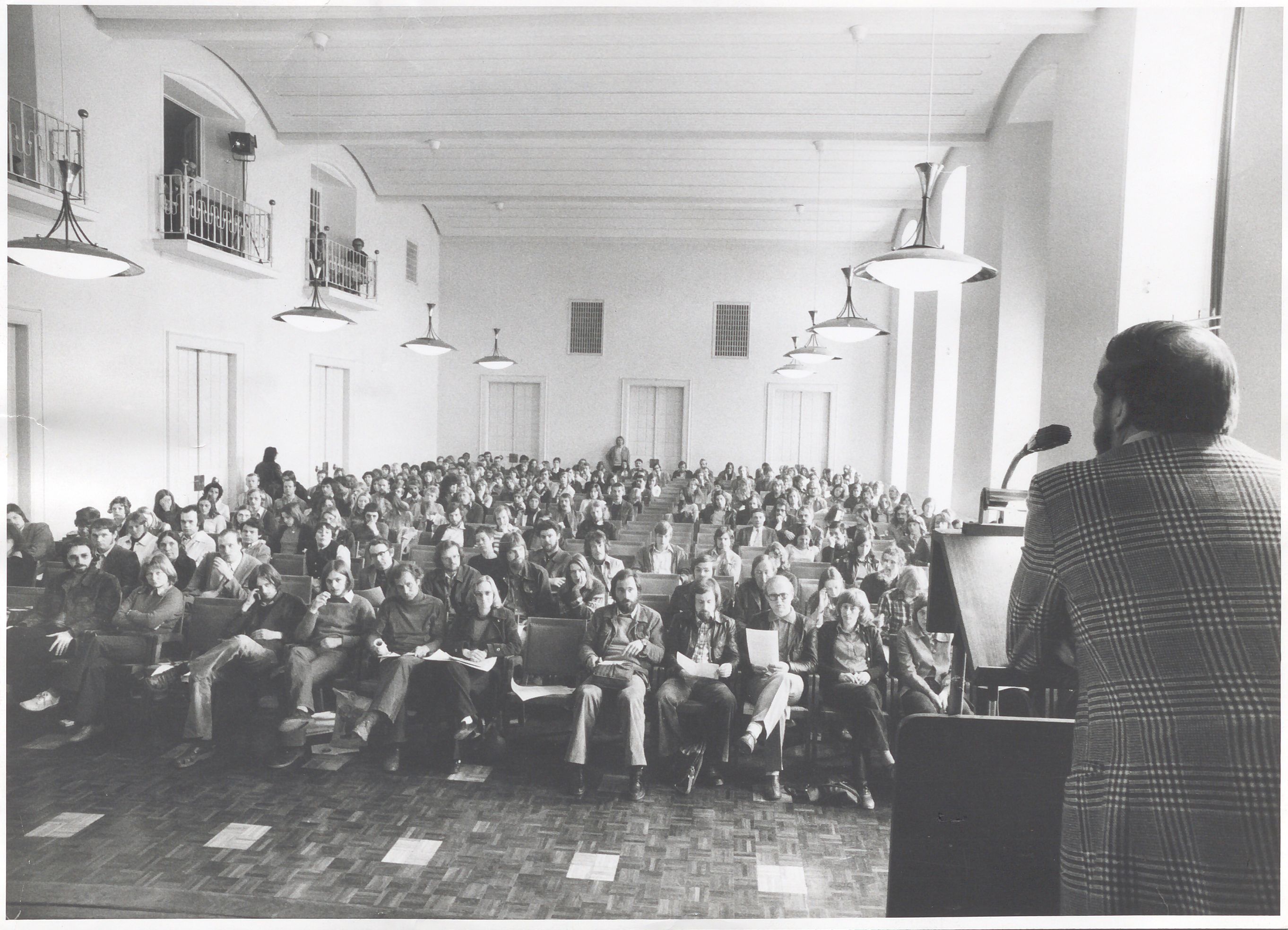 A black and white shot of a lecture in a large hall. In the foreground, a speaker stands at a lectern while many members of the audience sit in rows and look towards him. 