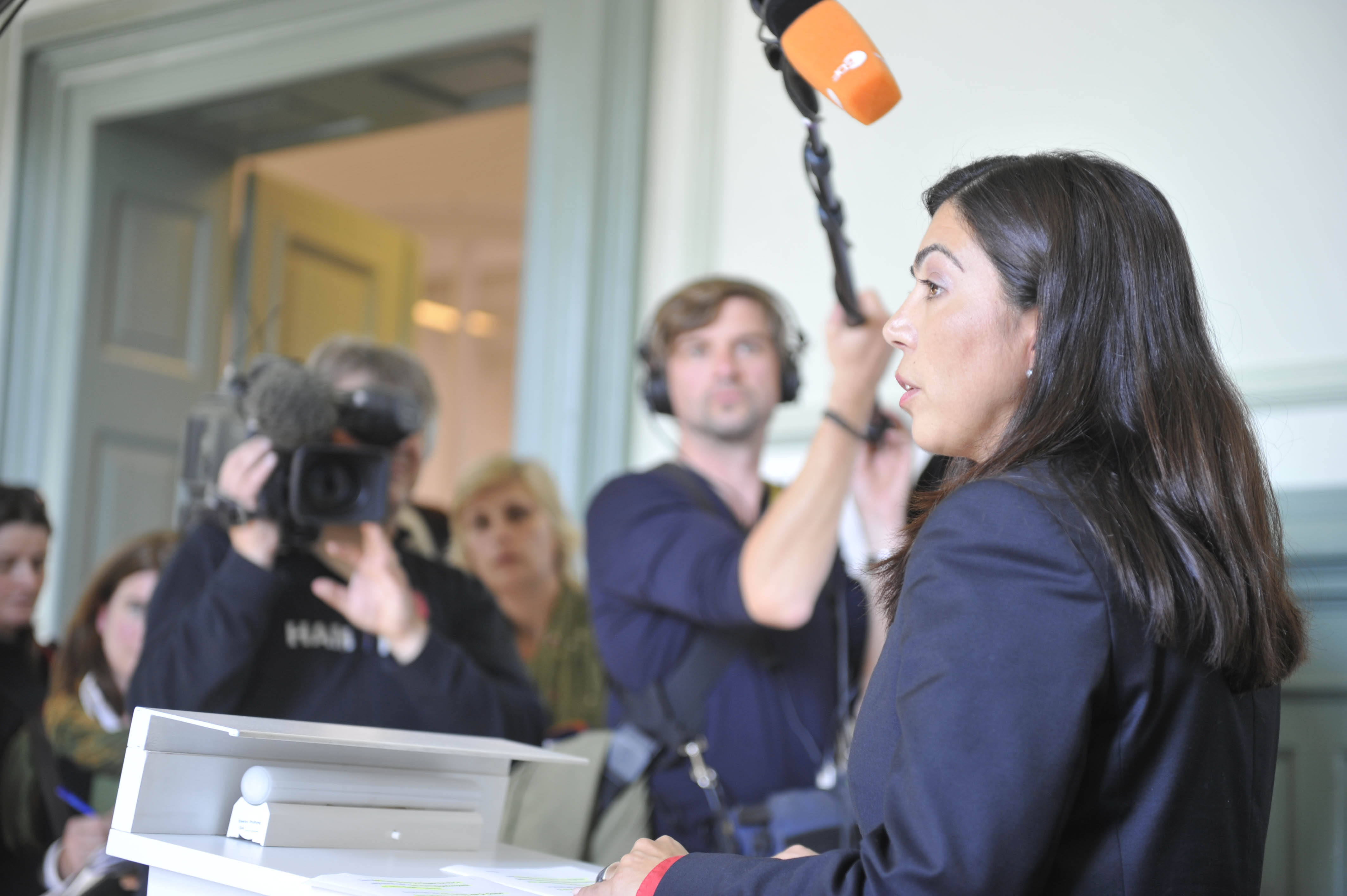 A woman in professional attire is speaking into a microphone during a press conference. In the background, several journalists and camera operators are seen, with one individual capturing video footage