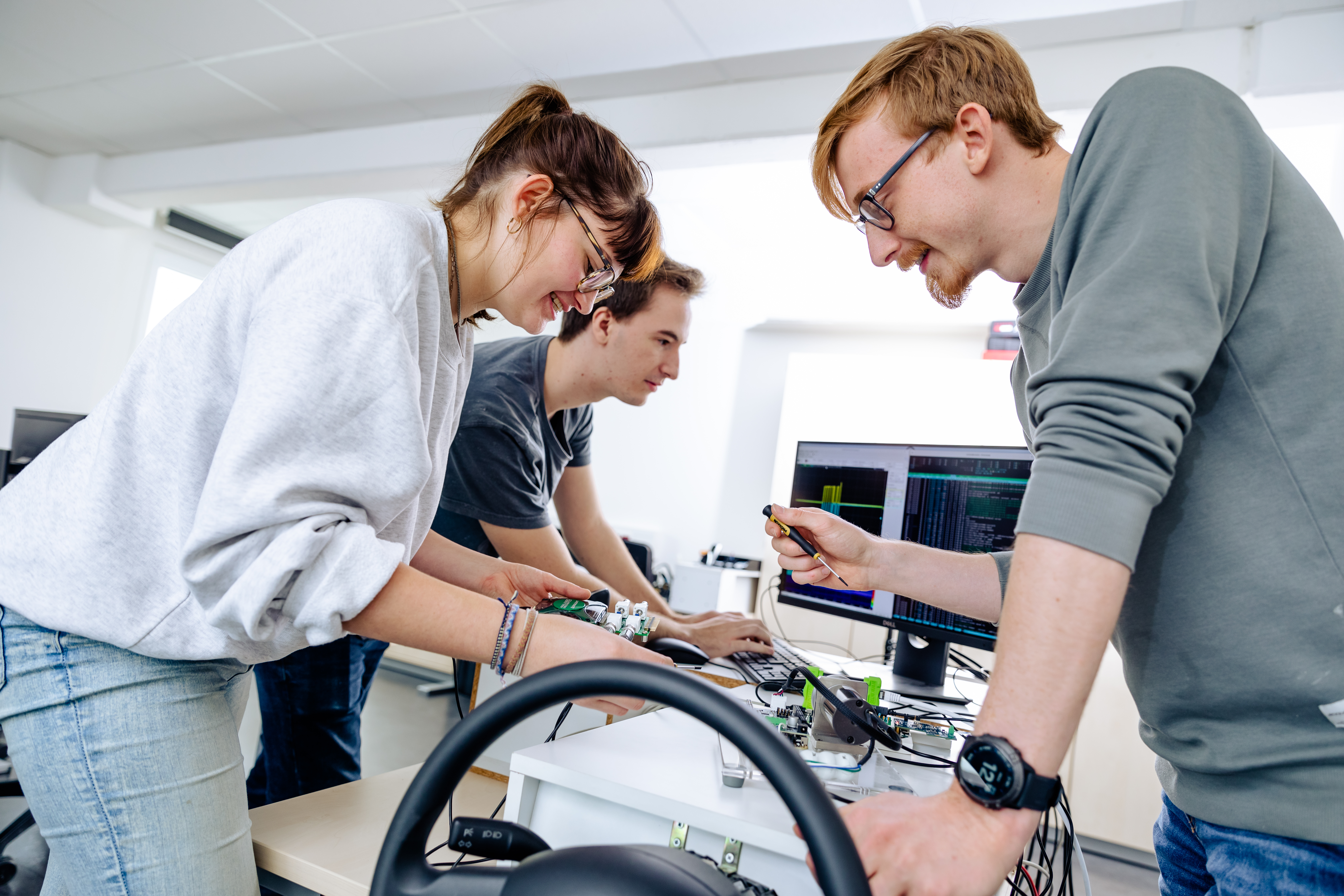 Three students are intently working on a technical project together. They are examining an electronic circuit and appear to be collaborating on a solution.